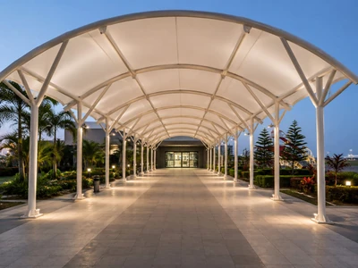 White arched fabric walkway canopy with steel posts and evening lighting leading to a building entrance