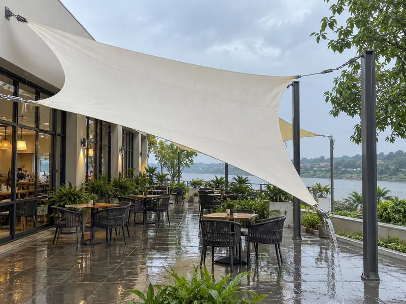 Steeply sloped ivory PVC shade sail over café seating with rainwater running off the low corner