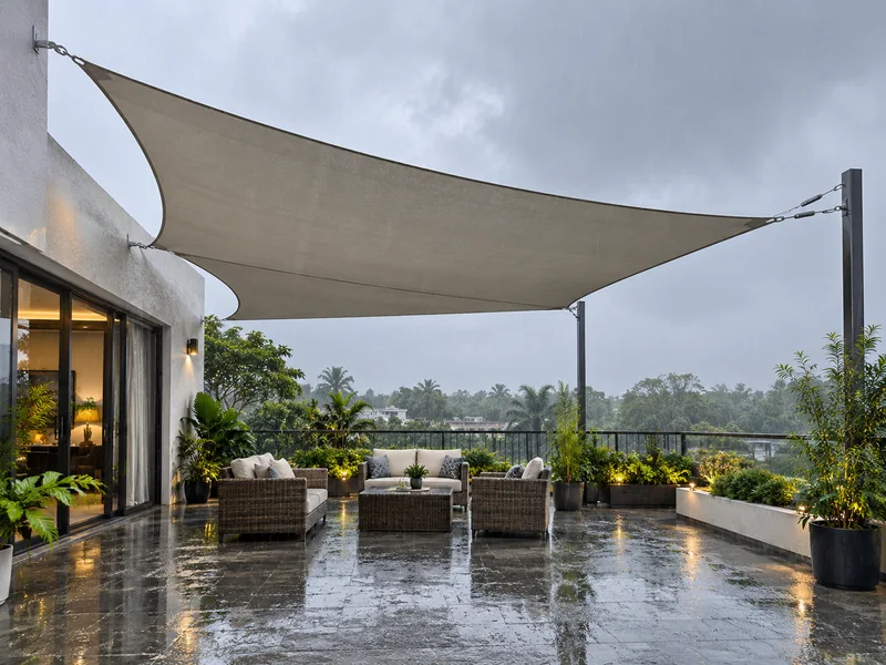 Waterproof shade sail over a terrace sitting area during monsoon rain, with dry chairs underneath
