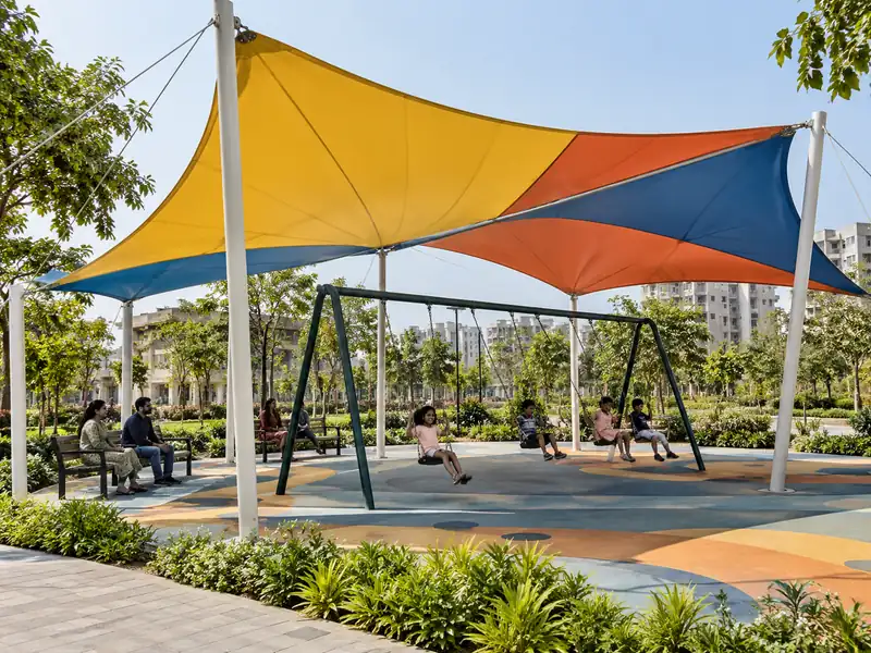 Colourful fabric canopy over swings in a public park with parents seated on benches in the shade