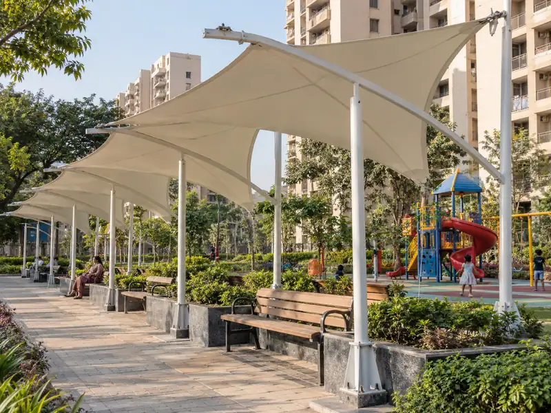 Row of membrane shade shelters over benches beside a children's play area in a housing society park