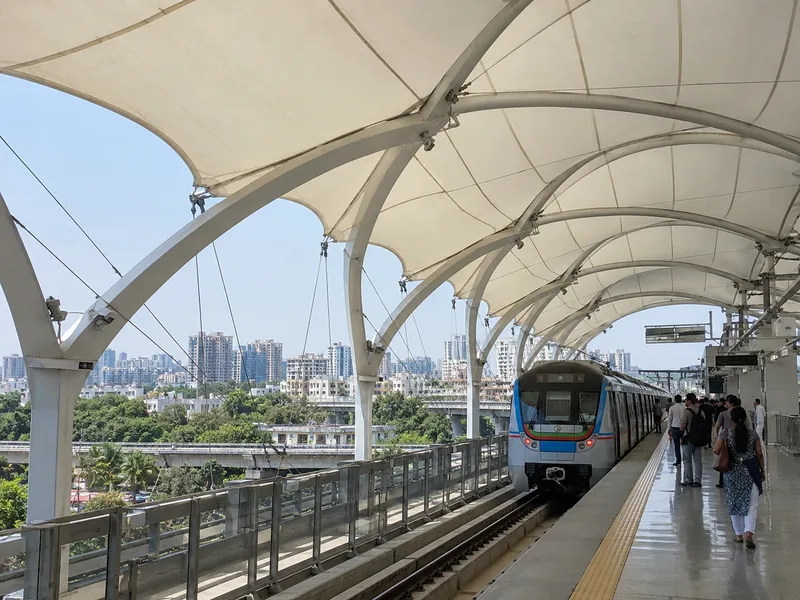 Tensile roof over an elevated metro platform with a train at the station and city views beyond